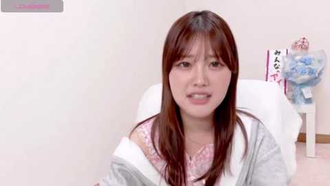 Video of an East Asian woman with long brown hair, wearing a pink lace bralette and gray hoodie, sitting on a white chair in a bright room with a floral arrangement in the background.