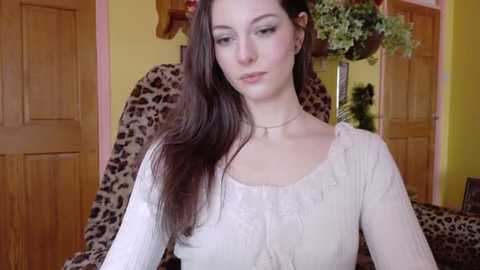 Video of a Caucasian woman with long dark hair, wearing a light-colored blouse, seated indoors, with wooden doors and a leopard print chair in the background.