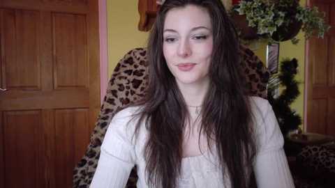 A video of a young woman with long, dark hair, wearing a white sweater, smiling in a cozy room with wooden doors, leopard-print chair, and a green plant.
