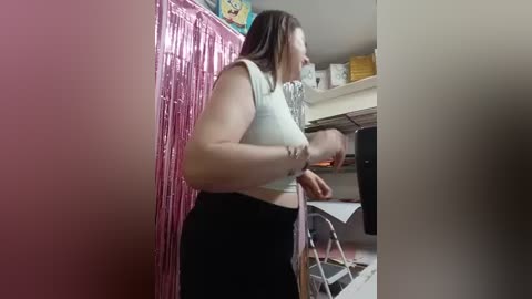 Video of a woman with fair skin and long brown hair, wearing a white crop top and black skirt, dancing in a cluttered kitchen with pink tinsel and white shelves.