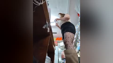 Video of a woman with fair skin and short brown hair, wearing a white tank top, black shorts, and patterned stockings, climbing a wooden ladder in a cluttered, brightly-lit room with shelves and containers.