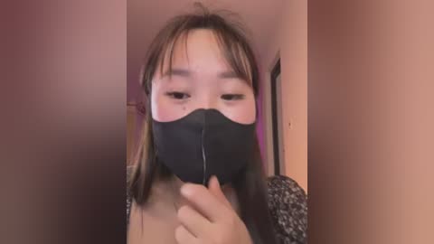 Video of an Asian woman with straight brown hair and light skin, wearing a black face mask, standing in a narrow corridor with peach-colored walls.