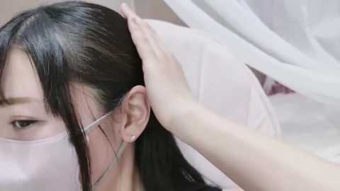 A video of an Asian woman with straight, dark hair, wearing a white face mask, lying on a white bed, having her head massaged by another person's hand.
