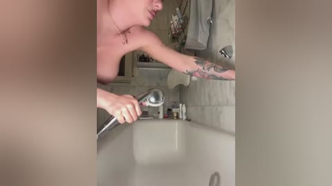 A video of a fair-skinned, tattooed woman, topless, washing her face in a white-tiled bathroom, holding a handheld shower head.