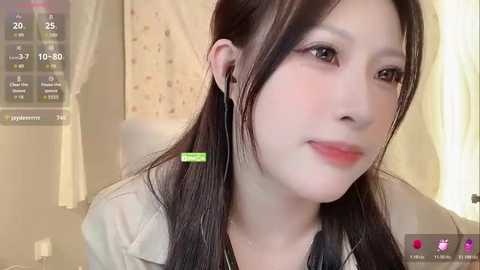 Video of a young East Asian woman with fair skin, long black hair, and soft features, wearing a beige jacket, with a green and black headphone. Background features a blurred indoor setting.