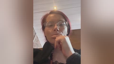 A video of an Asian woman with short red hair and glasses, resting her chin on her hand, in a dimly lit room with wooden ceiling panels.