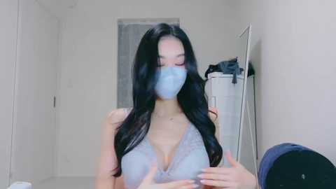 Media: Video of an Asian woman with long black hair, wearing a light blue lace bra, face mask, and standing in a white-tiled hallway.