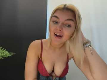 Video of a young blonde woman with fair skin, wearing a red lace bra and denim overalls, smiling and touching her hair. Background includes a dark wall and a green plant.