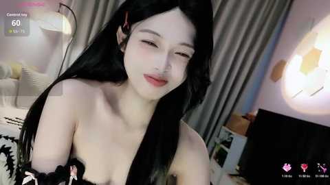 Video of an East Asian woman with long black hair, fair skin, and a slim physique, wearing a black off-shoulder top, smiling. Background features a modern living room with a lamp, curtains, and a sofa.