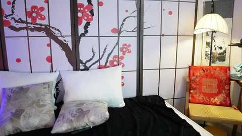 Video of a cozy bedroom featuring a black bed with white pillows and a grey-patterned throw. The background showcases a traditional Japanese screen with cherry blossom and red floral designs. A red fabric chair with gold embroidery is to the right.