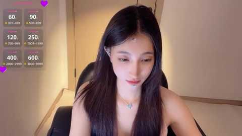 Video of an East Asian woman with long, straight black hair, fair skin, wearing a black top, sitting in a modern room with beige walls. A digital overlay displays heart icons and user stats.