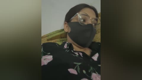 A video of a woman with medium brown skin and straight black hair, wearing a black floral blouse, clear glasses, and a black face mask. She sits on a green and yellow striped couch in a dimly lit room.