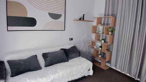 Video of a modern, minimalist living room with a white sofa, gray pillows, abstract wall art, wooden shelving unit, and light-colored curtains.