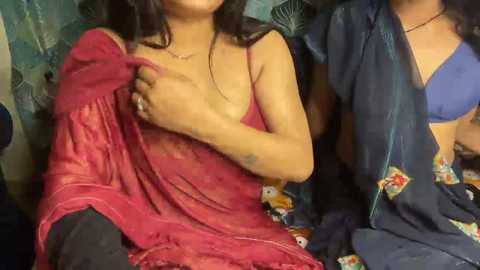 Media: Video of a South Asian woman with medium skin tone, long dark hair, wearing a sheer red saree, exposing her small breasts, sitting in a dimly lit room with colorful fabric and plants in the background.