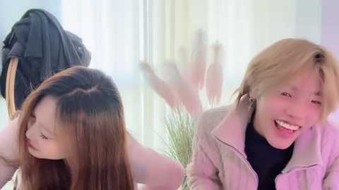 Video of two young East Asian women laughing, one with long straight brown hair, the other with short blonde hair, both in casual attire, sitting in a brightly lit room with a window.