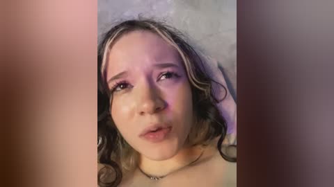 Media: A close-up video of a young woman with fair skin, wavy brown hair, and light makeup, lying on a white pillow, looking slightly distressed.