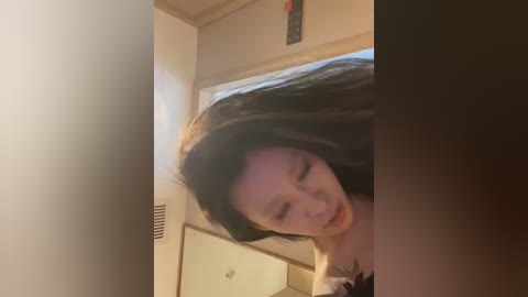 Media: Video of a young Asian woman with long black hair, wearing a black top, captured in a blurred motion, leaning against a beige wall in a dimly lit room.