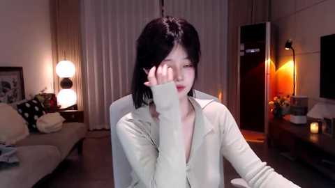 Video of an Asian woman with shoulder-length black hair, wearing a beige cardigan, covering her face with her right hand, sitting in a dimly lit living room.