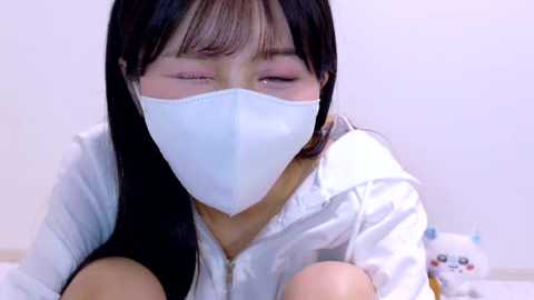 Media: A video of an East Asian woman with long black hair, wearing a white mask, pink eyeshadow, and a white shirt, with a soft, blurred background featuring a white teddy bear.