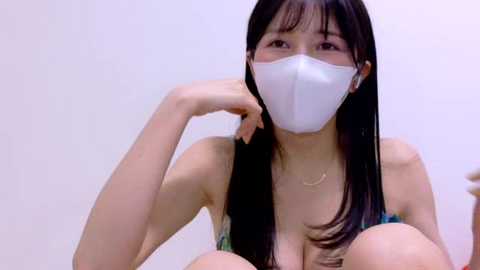 Video of an Asian woman with long black hair, wearing a white face mask, revealing cleavage in a low-cut green top.