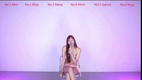Video of a nude Asian woman with long brown hair, sitting on a chair, holding a microphone, in a white room with \"No. 1 Miyay No. 2 Mina No. 3 Mana No. 4 Minami\" written in red text above.