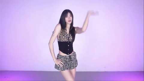 Media: Video of an Asian woman with long black hair, wearing a leopard-print top and skirt, posing against a white backdrop with purple lighting, striking a dynamic pose.