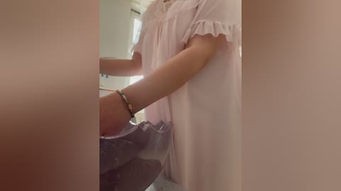 A video of a person in a light pink, ruffled blouse, holding a black garment. The background is blurred, showing a white wall and a window.