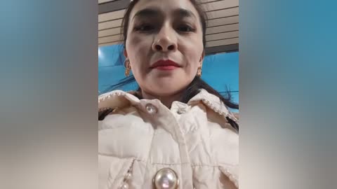 Video of a young woman with straight, dark hair, wearing a white puffy jacket with a silver button, smiling. Background features a clear blue sky and white roof.