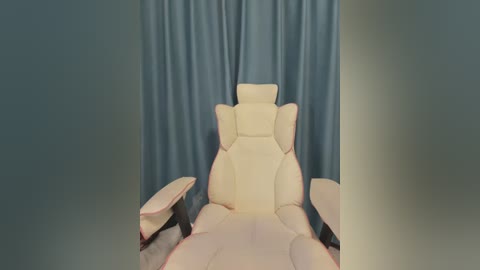 Media: Video of a white, modern ergonomic massage chair with high backrest and armrests, positioned against a backdrop of soft blue curtains. The scene is minimalist and serene, focusing on the chair's sleek, comfortable design.
