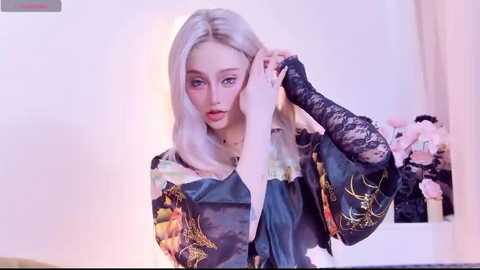 Video of a young woman with fair skin, platinum blonde hair, and red lips, wearing a black kimono adorned with floral patterns. She holds a lace glove against her face, in a softly lit room with a vase of pink roses.