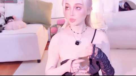 Video of a pale-skinned, platinum-blonde woman in black lace lingerie, sitting on a white couch with a green pillow. Background shows a cluttered living room with a pink blanket and books.