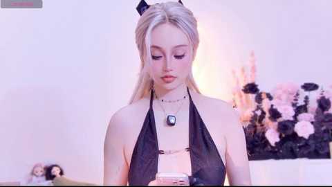 Video of a young, fair-skinned woman with platinum blonde hair in a halter top, looking down at a pink phone in a softly lit room with a floral arrangement in the background.