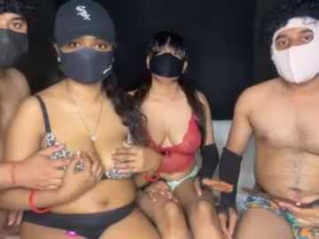 Video of four people wearing masks and colorful, revealing outfits. Two women, one with dark skin and a black bra, the other with lighter skin and a red bra, hold hands. Two men, one with a white mask, the other with a black mask, wear colorful briefs.