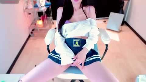 Media: Video of an East Asian woman with long black hair, wearing a white off-shoulder sweater and blue short skirt, sitting provocatively with legs spread, in a brightly lit, modern room.