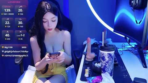 A video of an Asian woman with long black hair, wearing a yellow strapless dress, sitting in a dark room with a desk covered in sex toys and a large ceramic jar.