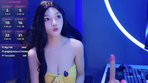 Video of an Asian woman with long black hair, wearing a yellow strapless dress, holding a large pink dildo, in a dimly lit, blue-themed room with a digital screen displaying statistics.