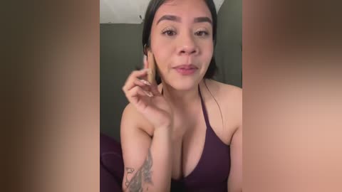 A video of a Latina woman with dark hair and medium skin tone, wearing a low-cut, dark purple top, sitting on a purple couch. She has a tattoo on her left arm. The background is a plain, dark wall.