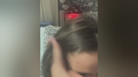 A blurry video showing a person with long brown hair, partially covered by a pink cloth, lying on a bed with white sheets. The background has a heart-patterned wall and a faint red heart emoji.