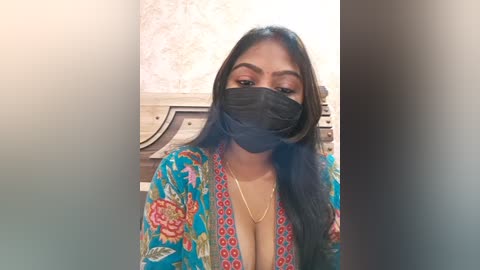 Video of a South Asian woman with long black hair, wearing a colorful floral blouse, black face mask, and gold necklace. She stands indoors with a wooden chair and patterned wallpaper in the background.