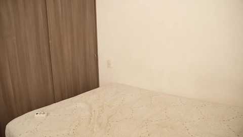 A video of a minimalist bedroom corner featuring a beige mattress on the floor, a brown wooden wardrobe, and a plain white wall, with a single electrical outlet visible.