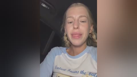 Video of a young woman with light skin, blonde shoulder-length hair, and a neutral expression, wearing a light blue and white long-sleeved shirt. She is seated in a dark vehicle with a blurred background.