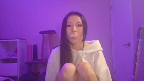 Video of an East Asian woman with long black hair, wearing a white hoodie, sitting on a floor, with a gauze pad taped over her mouth. The room has purple lighting, a cat tree, and shelves with books.