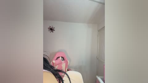 Video of a child lying on a bed in a small, white-walled room, wearing a pink jacket, with a black-haired doll beside them, a small star-shaped decoration on the wall, and a window visible on the right.