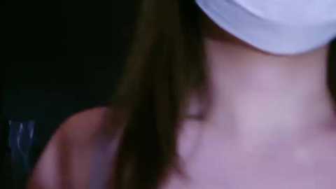 A close-up video of a woman with long, dark hair, wearing a white surgical mask, her face partially obscured, in a dimly lit setting.