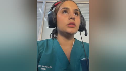 A video of a young woman with light brown skin, wearing headphones and a teal scrub top with a name tag. She has a neutral expression and is standing in a clinical setting with white walls and a window.