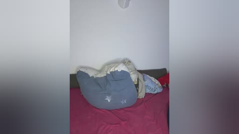 A video of a child lying on a bed with a pink sheet, wearing a blue shirt with white stars, under a white blanket, in a dimly lit room.