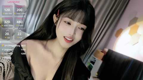 Video of an East Asian woman with long black hair, wearing a low-cut black top, smiling in a dimly-lit room with a lamp and a TV screen showing viewer counts.