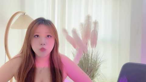Video of a young Asian woman with long brown hair, fair skin, and light makeup. She is topless, wearing a pinkish glow. Background includes a floor lamp, tall pampas grass, and sheer white curtains.