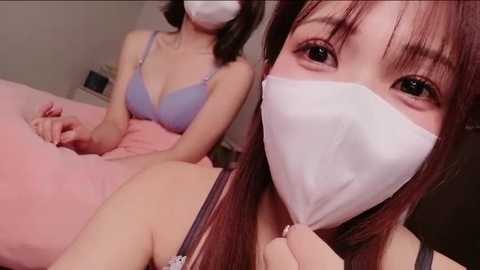 Video of two East Asian women wearing masks, sitting on a bed. One wears a light blue bra, the other a white mask. Pink sheets in the background.