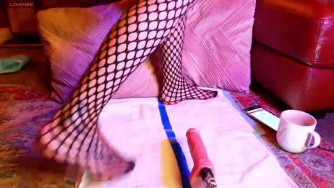 Video of a person with black fishnet stockings and red heels on a carpeted floor, near a white mug, a smartphone, and a red vibrator.
